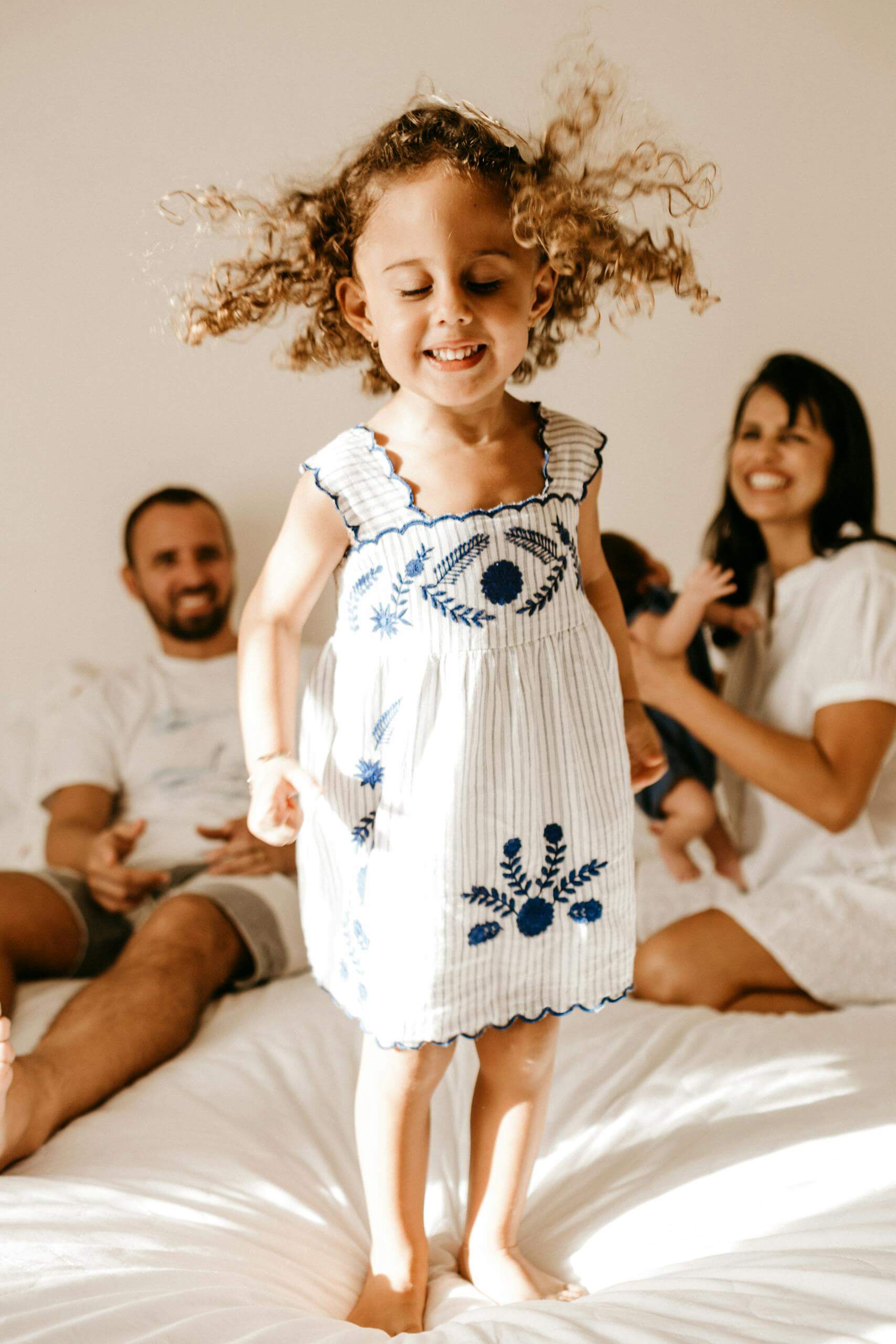 Adorable little girl with curly hair in light dress jumping on bed with closed eyes near happy young parents and newborn baby. Reach out today to begin with parenting support for moms, dads, parents, and to better the connection with your child. 