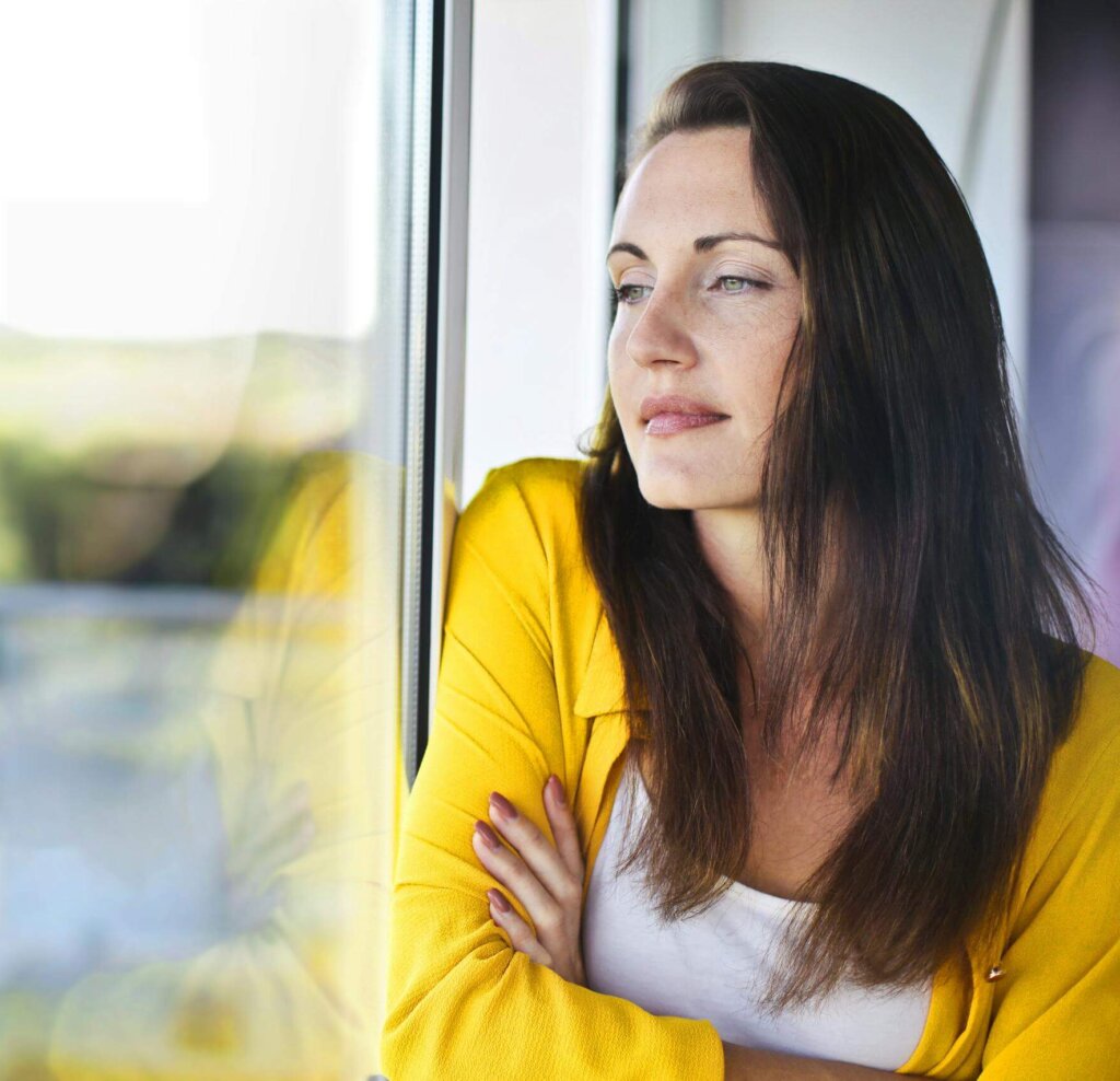 Woman sitting by a window looking thoughtful, representing the quiet overwhelm many moms in Matthews, NC experience before starting therapy